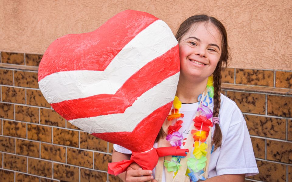 Ein Mädchen lächelt fröhlich und hält ein großes rotes Herz mit weißen Streifen in den Armen.