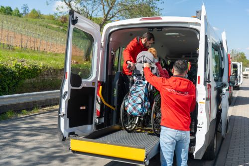 Das Bild zeigt den Einstieg einer Person im Rollstuhl in ein weißes Fahrzeug des Fahrdienstes durch eine barrierefreie Rampe mit Unterstützung der Mitarbeitenden.